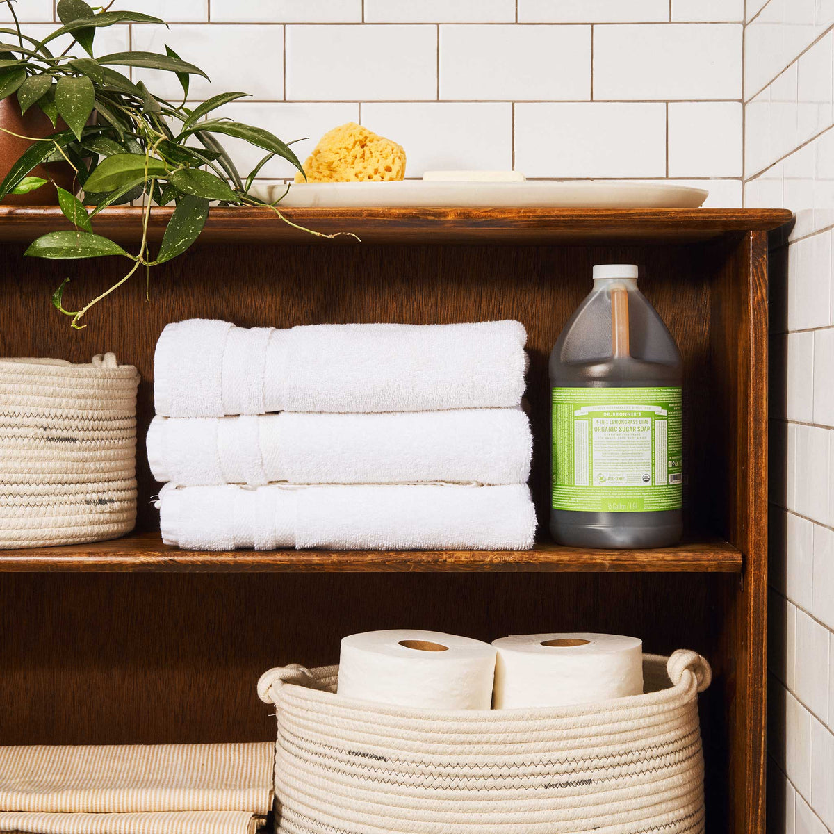 A wooden shelf holds a bottle of Dr. Bronners Organic Sugar Soap with white labels, surrounded by neatly folded white towels, a sponge, a plant, and rolls of toilet paper in a woven basket, set against tiled walls [1/2 Gallon]