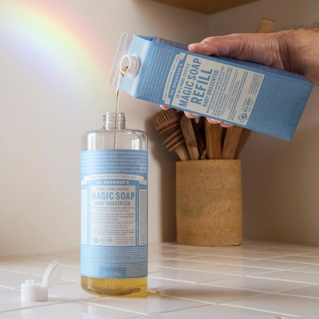 A bottle of Dr. Bronners baby unscented castile soap being refilled on a kitchen counter, with a rainbow reflection on the wall. A container with utensils is in the background [32 oz]