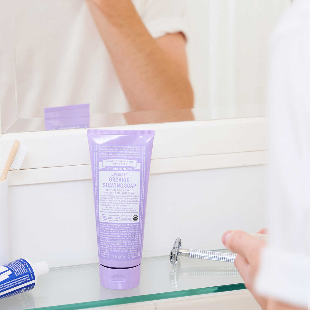 A lavender tube labeled Dr. Bronners sits on a bathroom sink beside a toothbrush and a razor, with a person reflected in the mirror above, suggesting a morning routine [7 oz]