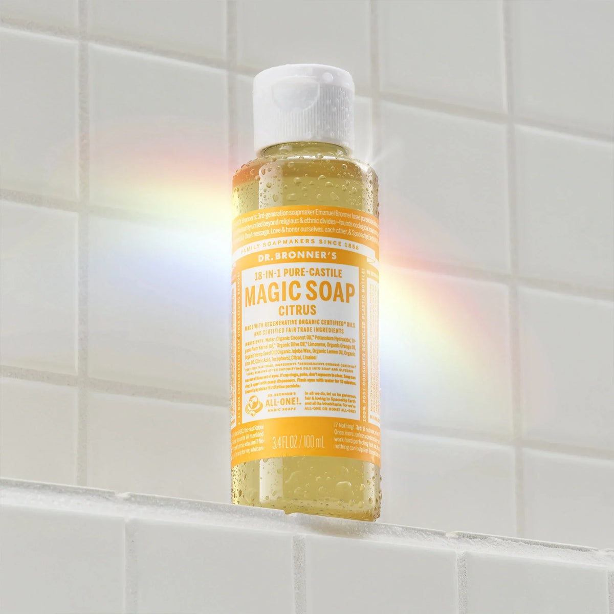 A bottle of Dr. Bronners Citrus Pure Castile Magic Soap sits on a white tiled shelf, illuminated by a rainbow light. Text reads 18 IN 1 Pure Castile MAGIC SOAP Citrus 3 FL OZ 89 mL [3.4 oz]