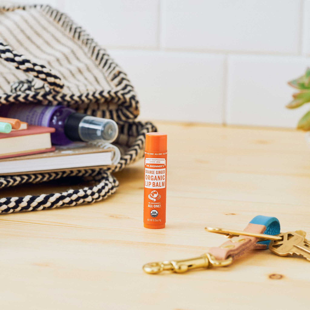 A Dr. Bronners lip balm stands upright on a wooden surface with a patterned bag nearby, partially open to reveal a book and a small bottle, beside a set of keys [.15 oz]