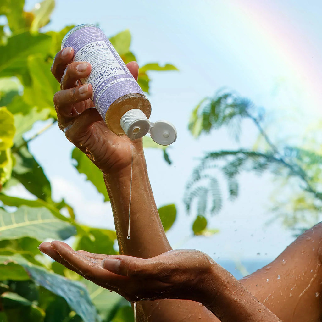 A person squeezes liquid from a bottle of Dr. Bronners lavender pure castile soap into their palm, surrounded by lush greenery and a faint rainbow. Text on bottle DR. BRONNERS Pure Castile Liquid Soap. Keywords castile soap, organic castile soap, vegan soap, Dr. Bronners uses [8 oz]