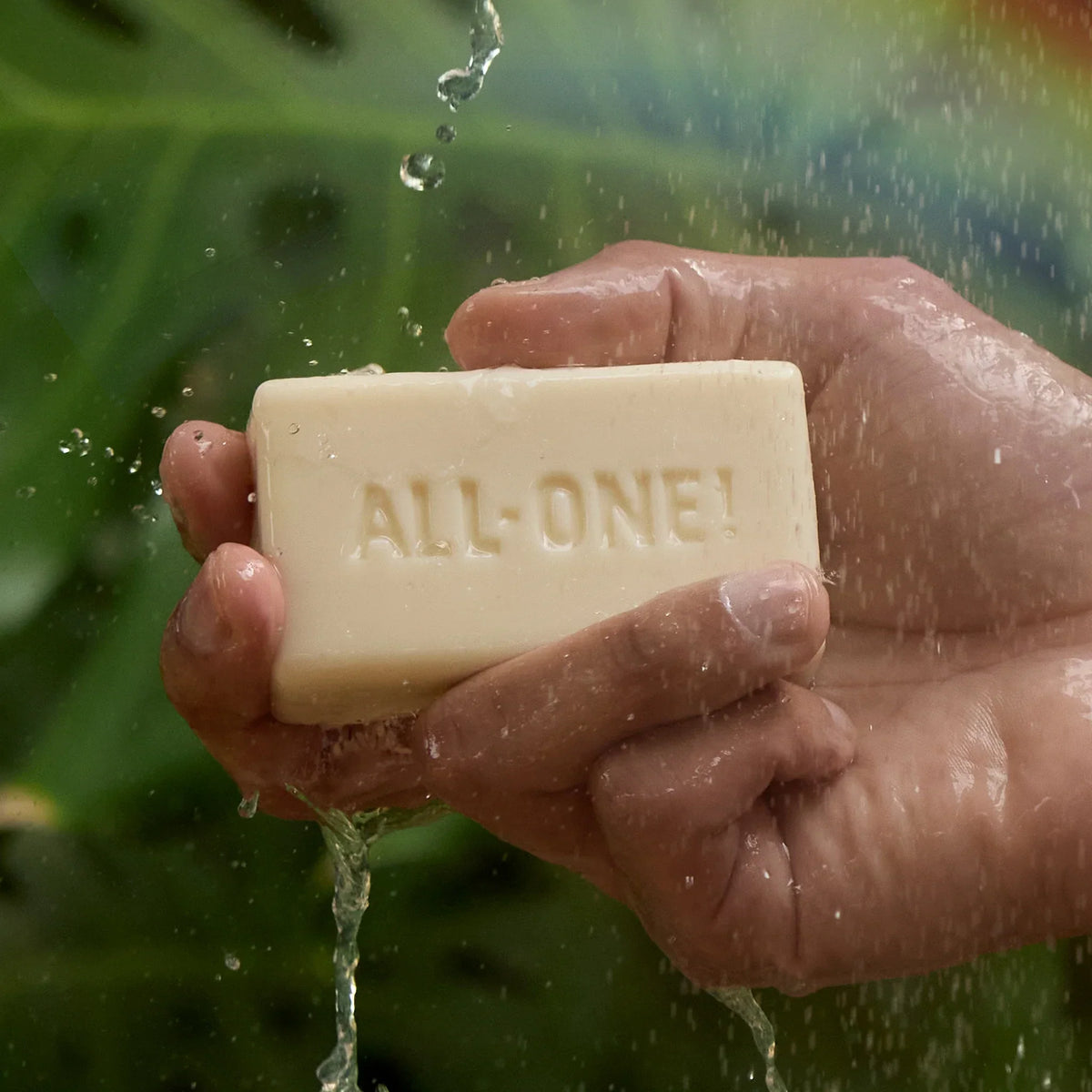 A hand holds a bar of soap embossed with ALL ONE beneath flowing water, set against a leafy, green background. This suggests a natural, eco friendly context ideal for organic soap use [5 oz]