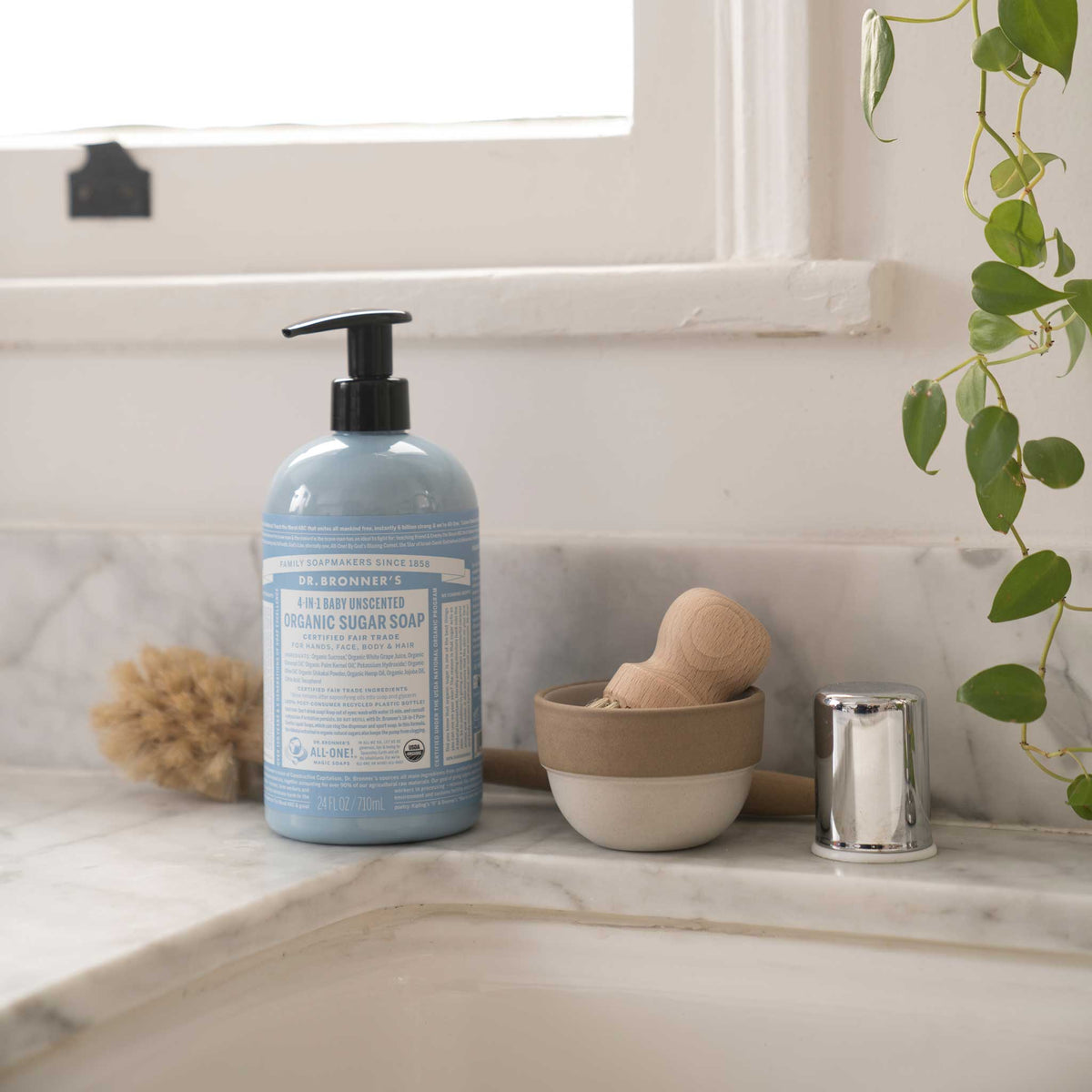 A bottle of Dr. Bronners liquid soap stands on a marble countertop beside grooming tools. The sunlit window and green plant enhance an eco friendly bathroom ambiance. Text on the bottle includes product details [24 oz]