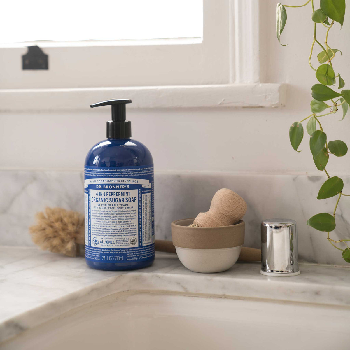 Dr. Bronners 4 in 1 Peppermint Organic Sugar Soap bottle sits on a marble counter by a window, surrounded by a wooden brush, ceramic bowl, and green plant, highlighting an eco friendly bathroom setting [24 oz]