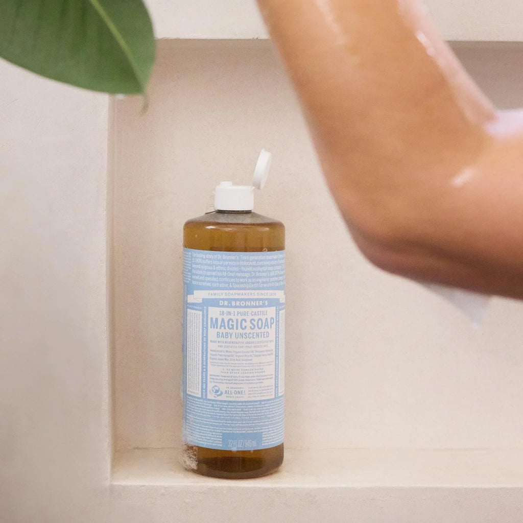 A bottle of Dr. Bronners Pure Castile Baby Unscented Soap stands in a shower niche with an open lid, next to a partially visible person washing against a white tile backdrop [32 oz]