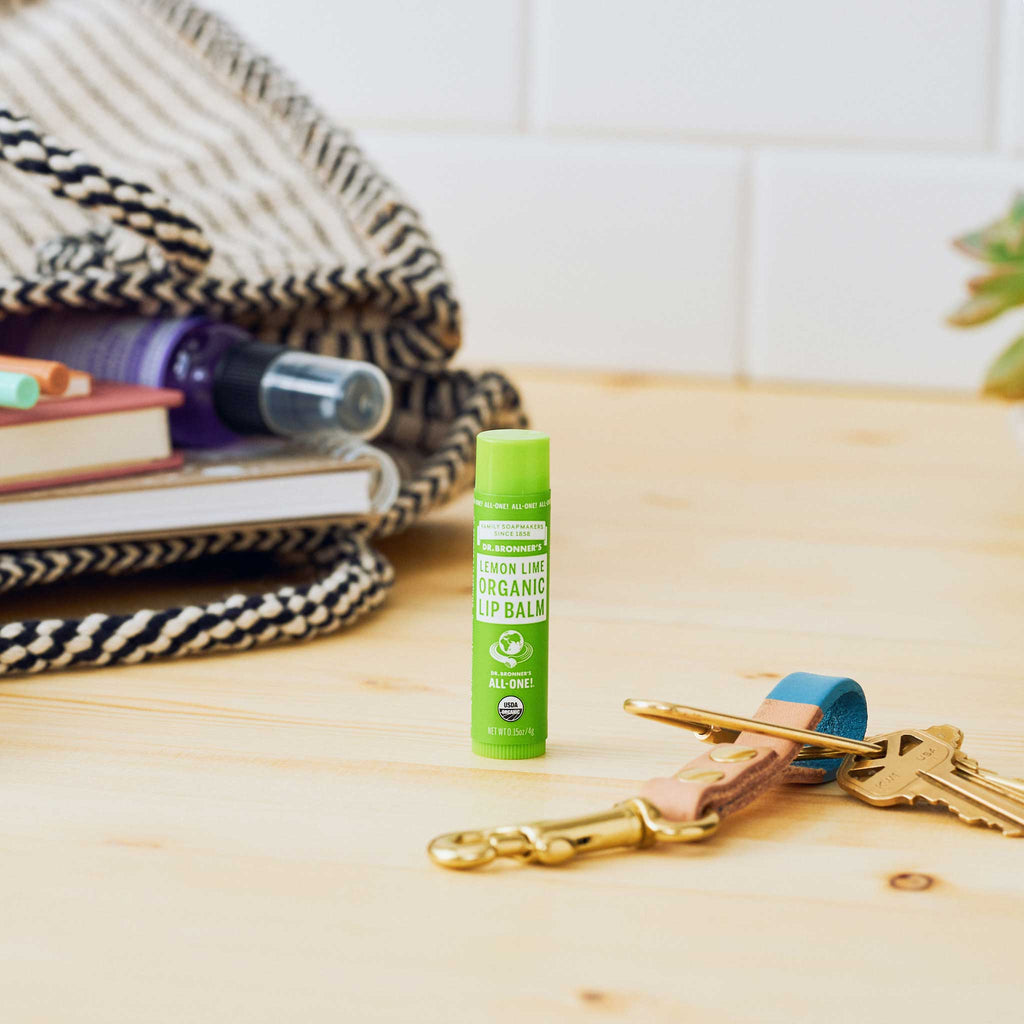 A lemon lime organic lip balm stands upright on a wooden surface beside a set of keys with colorful keychains. In the background, a patterned blanket and books create a cozy atmosphere [.15 oz]