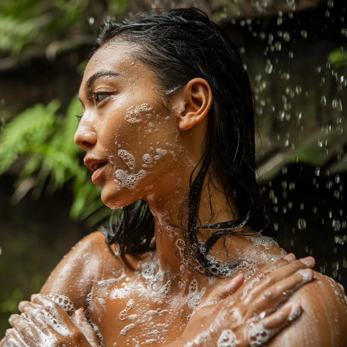 A person gently applies soap lather while showering outdoors, surrounded by lush greenery and cascading water, creating a natural and refreshing  bathing environment [2 oz, 3.4 oz, 4 oz, 8 oz, 16 oz, 32 oz, 1/2 Gallon, 1 Gallon]