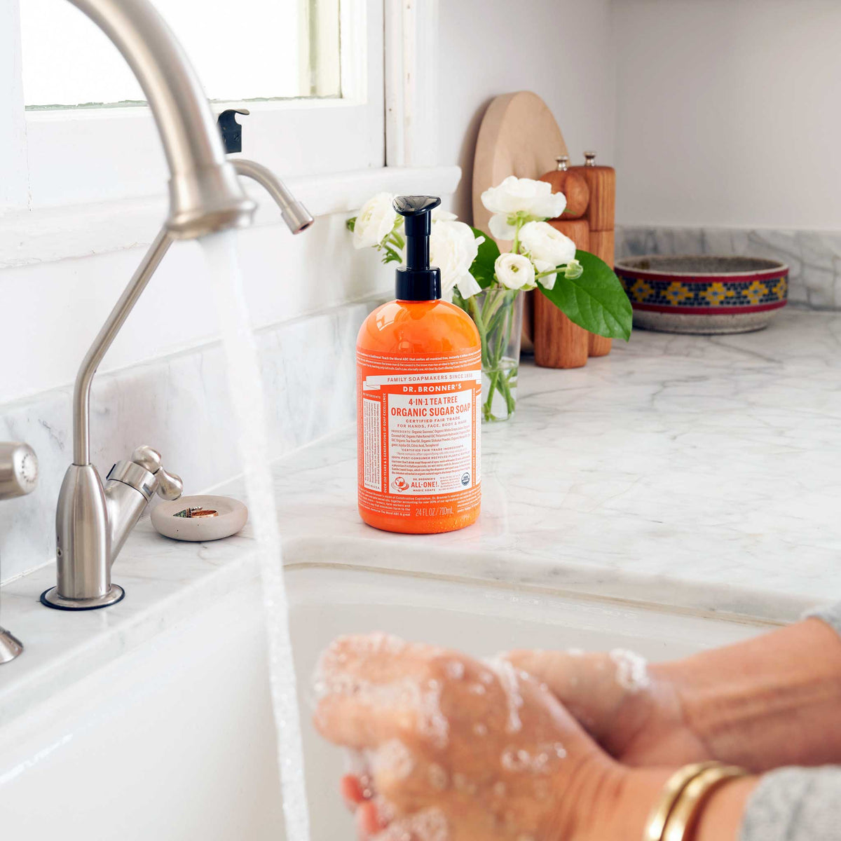 Dr. Bronners Organic Sugar Soap bottle sits on a marble counter beside a running faucet. Hands lather under the water. Nearby, flowers and kitchen items create a cozy setting [24 oz]