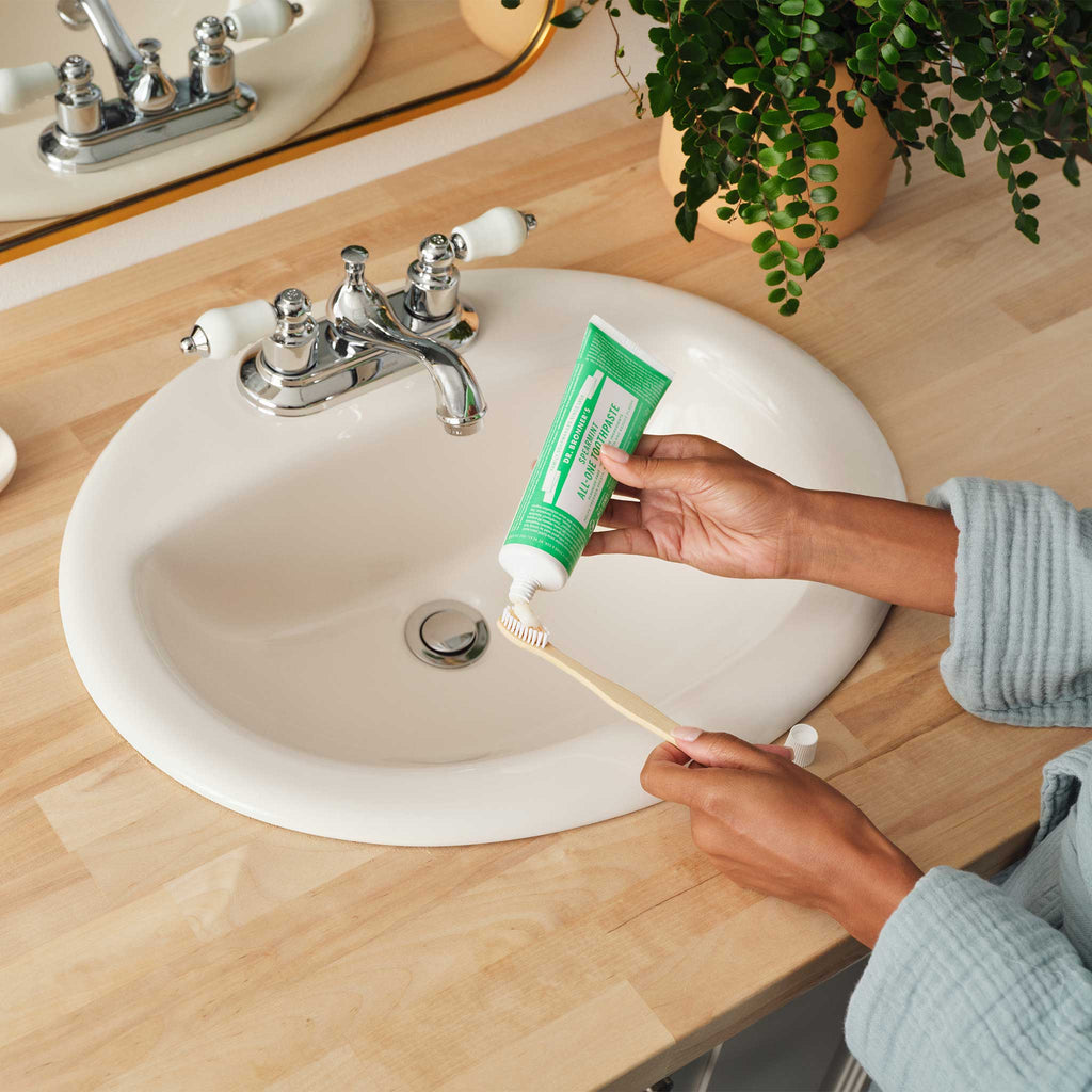 A hand squeezes Dr. Bronners peppermint toothpaste onto a bamboo toothbrush over a white sink. The setting features a wooden countertop and a potted plant, creating a natural, eco friendly bathroom scene [5 oz]