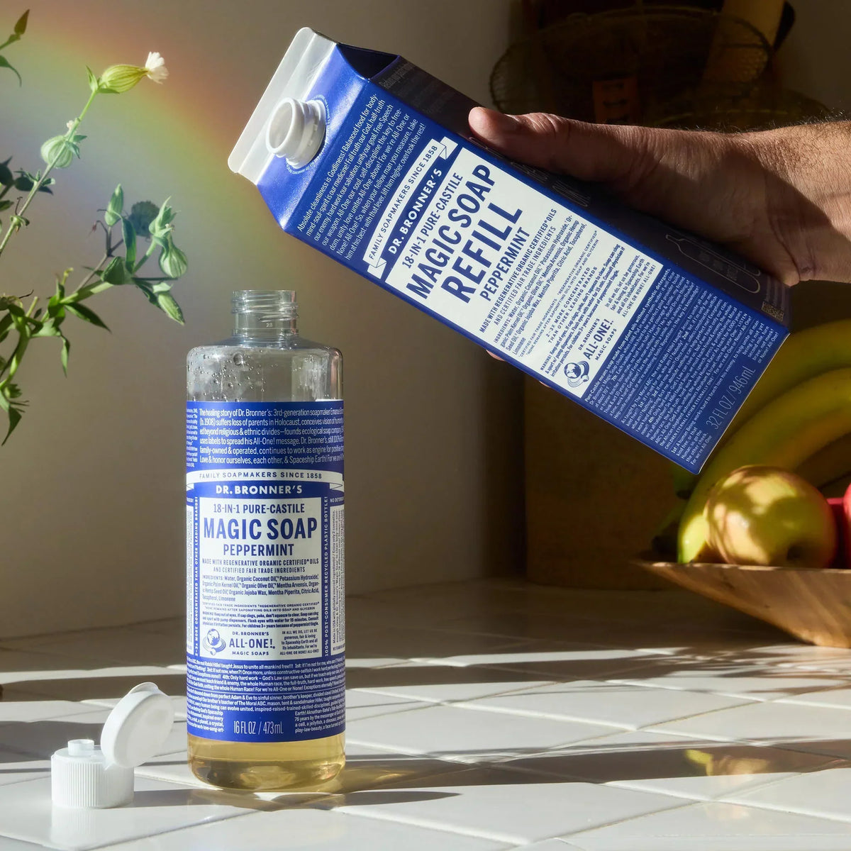 A hand pours Dr. Bronners Magic Soap Peppermint Refill into a bottle on a sunlit kitchen counter with a rainbow and fruit bowl in the background. DR. BRONNERS 18 IN 1 PURE CASTILE MAGIC SOAP PEPPERMINT MAGIC SOAP REFILL [32 oz]