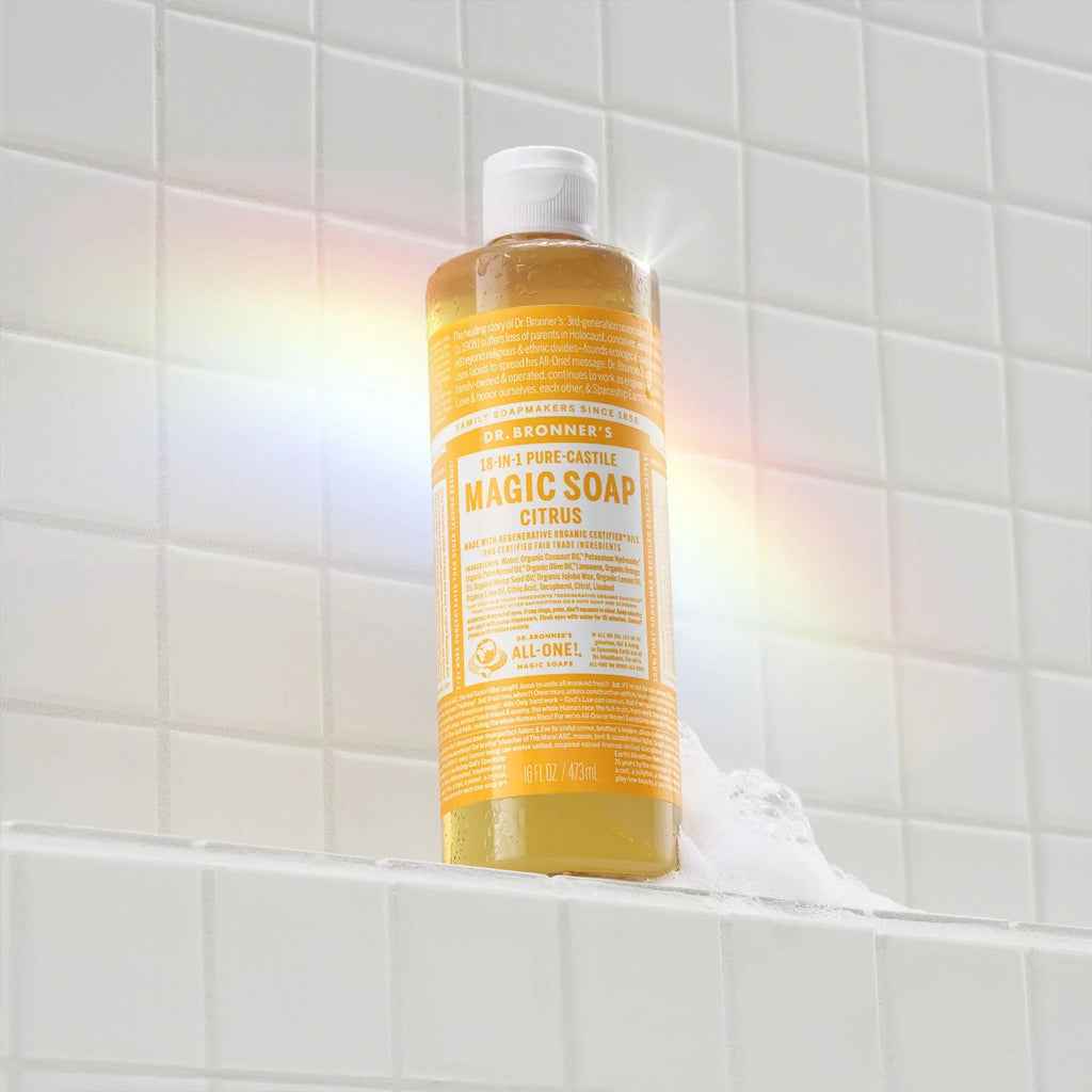 A bottle of Dr. Bronners 18 in 1 Pure Castile Magic Soap, Citrus, stands on a tiled ledge with soap suds. Rainbow light effects illuminate the scene, creating an eco friendly ambiance [16 oz]
