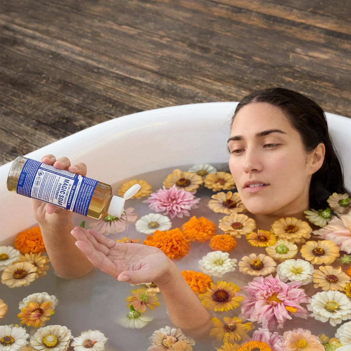 A woman reclines in a bathtub filled with water and colorful flowers, pouring Dr. Bronners soap into her hand, set against a wooden floor backdrop [8 oz]