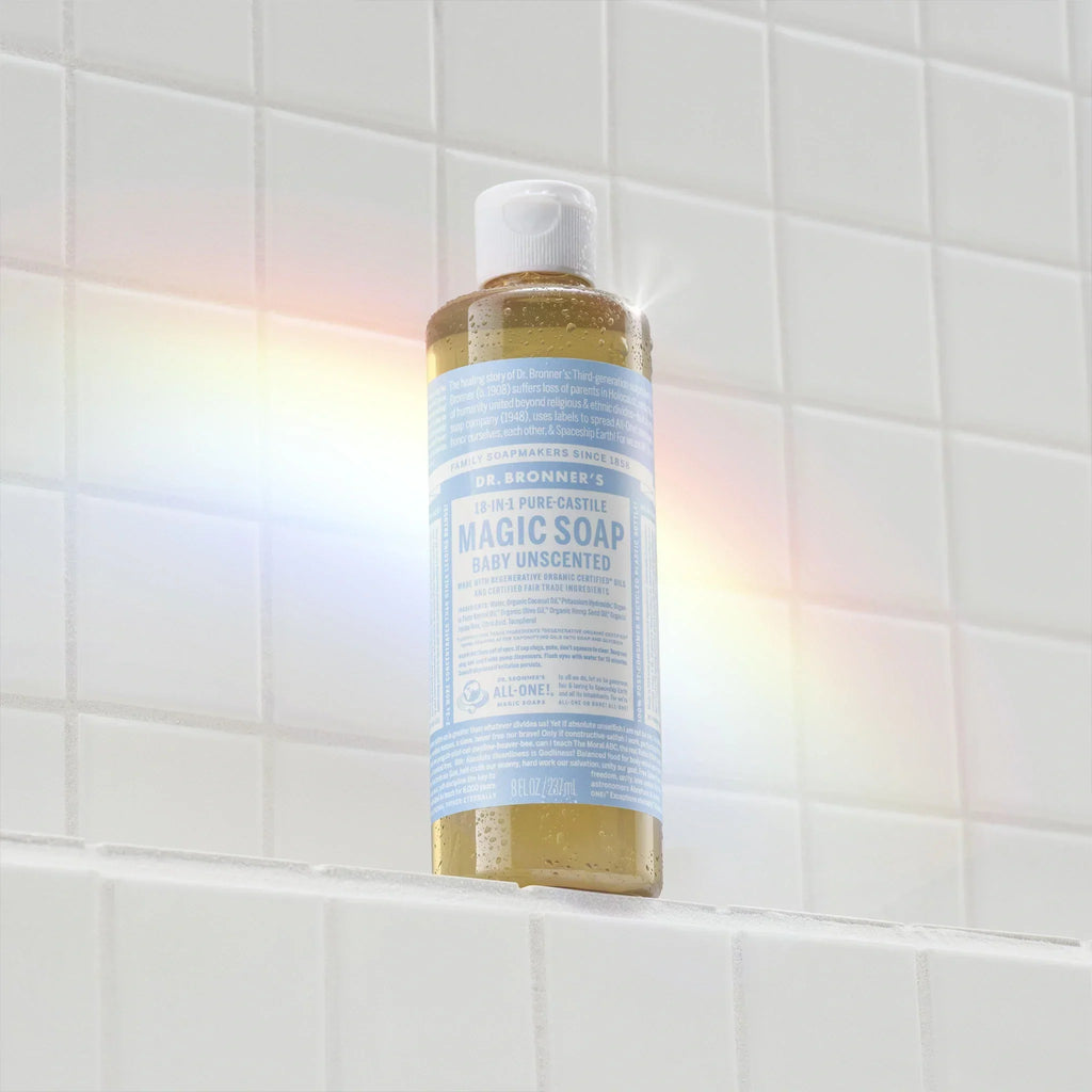 A bottle of Dr. Bronners 18 in 1 Pure Castile Magic Soap, Baby Unscented, sits on a white tiled surface, surrounded by a rainbow reflection. The label is detailed with extensive text [8 oz]