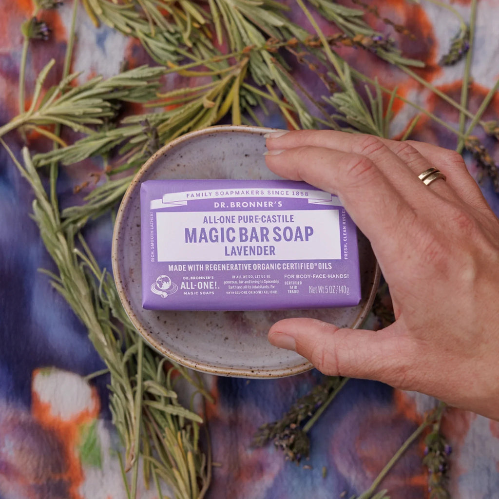 A hand holds a Dr. Bronners Lavender Magic Bar Soap on a ceramic dish, surrounded by lavender sprigs. FAMILY SOAPMAKERS SINCE 1858, DR. BRONNERS, ALL ONE PURE CASTILE MAGIC BAR SOAP LAVENDER, MADE WITH REGENERATIVE ORGANIC CERTIFIED FAIR TRADE OILS [5 oz]