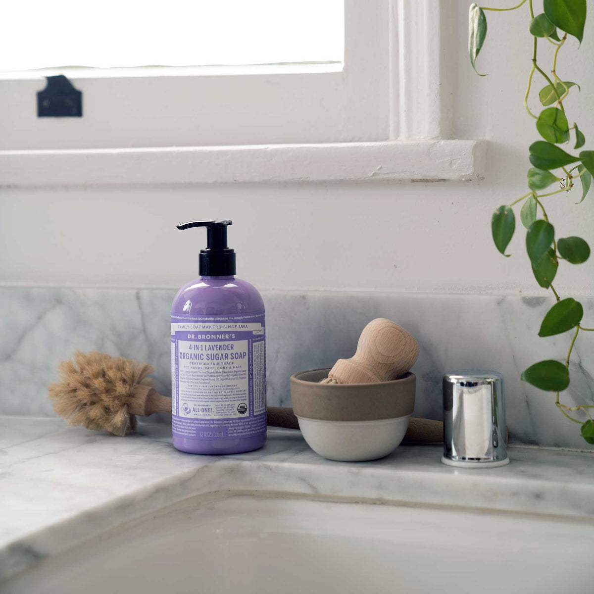 A pump bottle of Dr. Bronners lavender liquid castile soap sits on a bathroom countertop next to a sponge, dish, and greenery near a window, suggesting an eco friendly setting [12 oz]