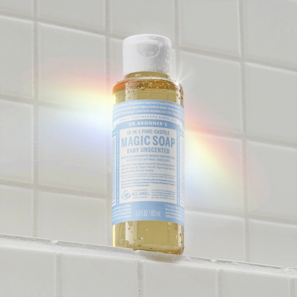 Dr. Bronners 3 in 1 Pure Castile Magic Soap, Baby Unscented version, sits on a white tiled surface, with a light rainbow reflection on the left, highlighting its eco friendly packaging [3.4 oz]