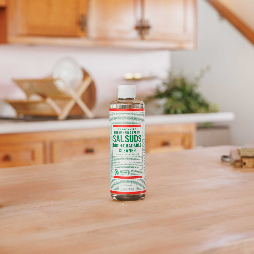 A bottle of Dr. Bronners peppermint castile soap sits on a wooden kitchen countertop, surrounded by a warm, rustic kitchen setting with wooden cabinets and greenery in the background [16 oz]