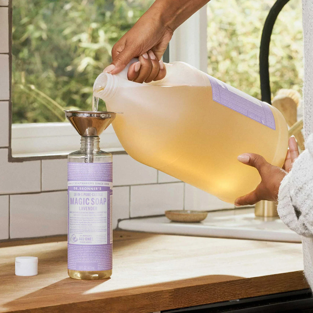 A lavender labeled bottle being refilled with liquid castile soap from a large jug using a funnel, set on a sunlit kitchen counter near a window overlooking greenery [16 oz]