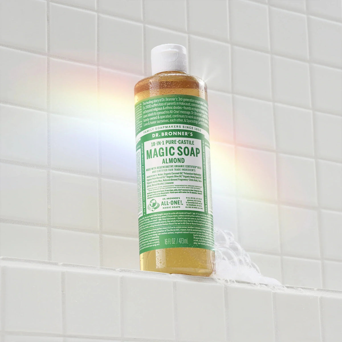 A bottle of Dr. Bronners eucalyptus castile soap stands on a white tiled shower ledge, with a soapy trail beneath it. A rainbow reflection appears on the tiles [16 oz]