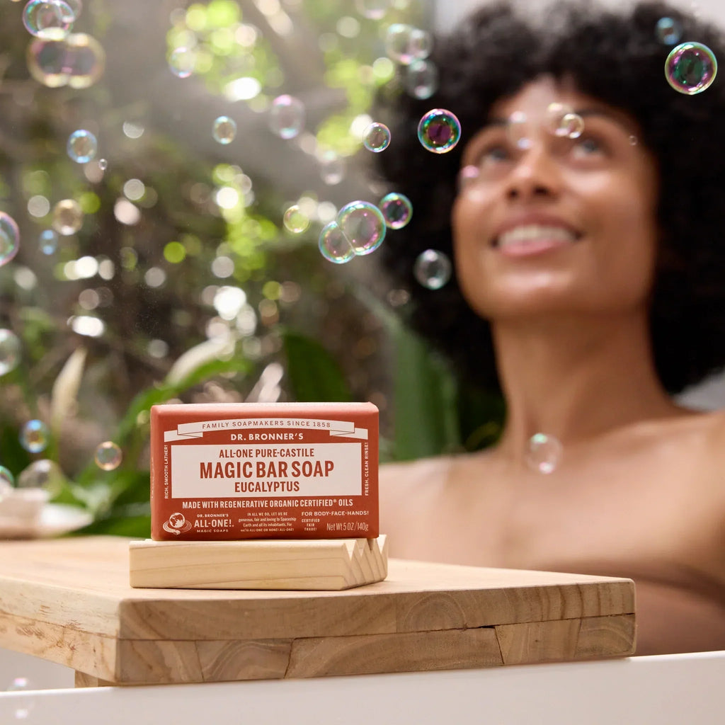 A bar of Dr. Bronners rose castile soap sits on a wooden ledge, surrounded by floating bubbles, while a person enjoys a bath in a plant filled setting [5 oz]