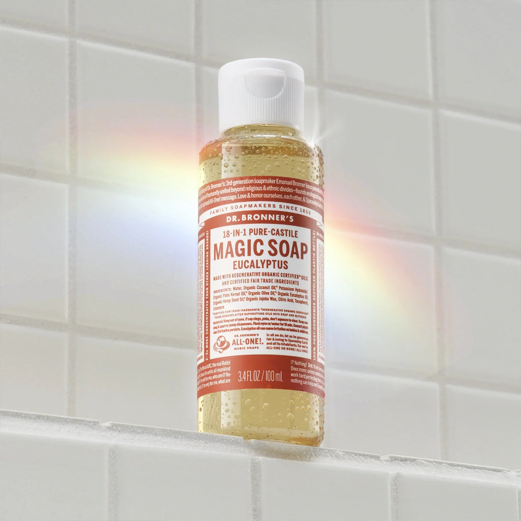 A bottle of Dr. Bronners 18 in 1 Pure Castile Magic Soap Eucalyptus sits on a white tiled shelf, with a rainbow reflection on the surface. Text includes DR. BRONNERS, 18 IN 1 PURE CASTILE, MAGIC SOAP, EUCALYPTUS, ALL ONE, 3.4 FL OZ 100 ml [3.4 oz]