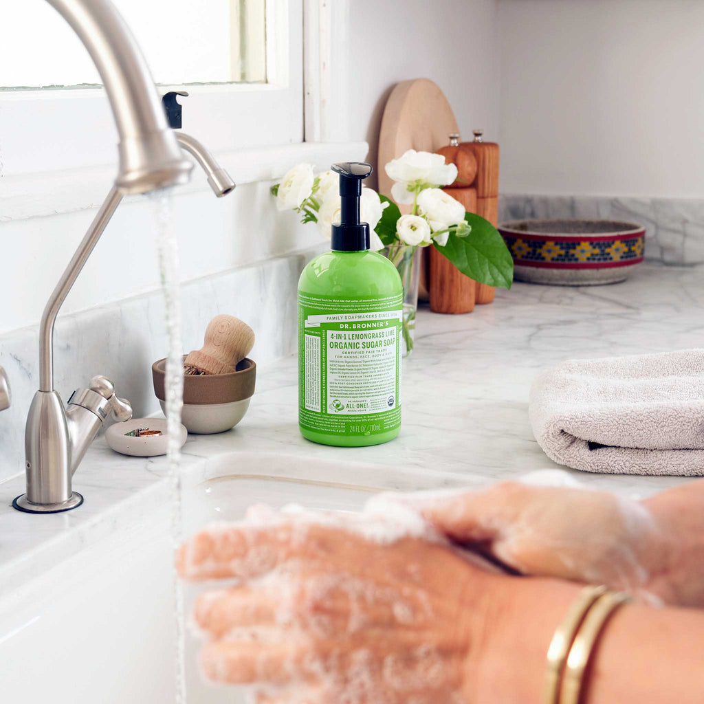 Hands lather with suds under running water from a silver faucet on a marble countertop. Nearby, a green bottle of Dr. Bronners Organic Sugar Soap rests beside a rolled towel and potted flowers [24 oz]