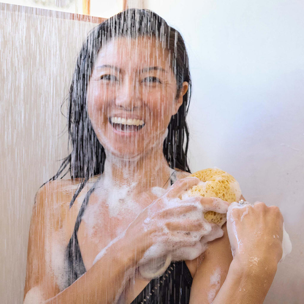 A person smiles while showering, using a sponge to apply soap on their shoulder. Water cascades down in a well lit bathroom, showcasing joyful self care [2 oz, 3.4 oz, 4 oz, 8 oz, 16 oz, 32 oz, 1 Gallon]