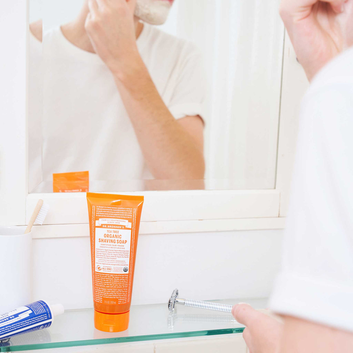 A person shaves in a bathroom with a mirror, using a tube of Dr. Bronners shaving soap. The orange tube stands on a glass shelf next to shaving essentials [7 oz]