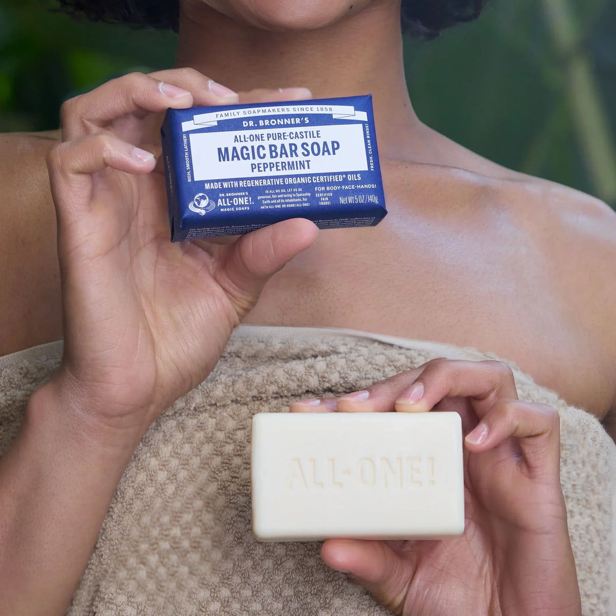 A person holds a bar of Dr. Bronners Baby Unscented Pure Castile soap, wrapped in blue packaging with white text, standing against a leafy background, emphasizing natural and vegan soap benefits [5 oz]