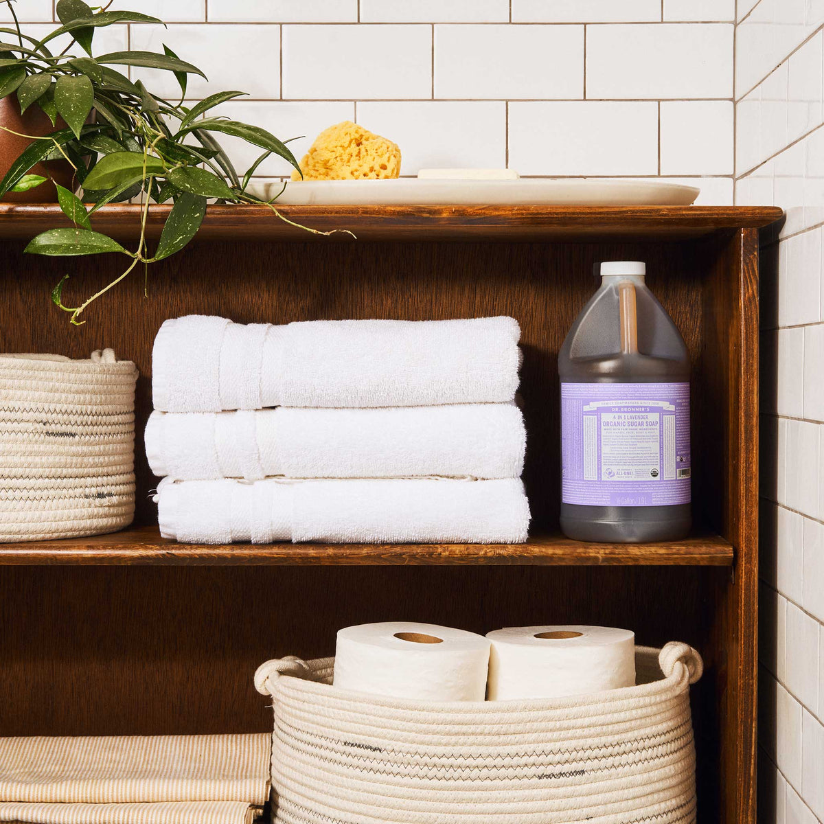 A wooden shelf in a tiled bathroom holds neatly stacked white towels, a bottle of Dr. Bronners lavender castile soap, toilet paper in baskets, a plant, and a sponge on top [1/2 Gallon]