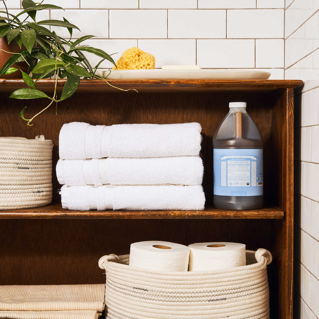 A bottle of Dr. Bronners liquid castile soap with a blue label sits on a wooden shelf. Surrounding it are neatly stacked white towels, a potted plant, and white tiles [1/2 Gallon]