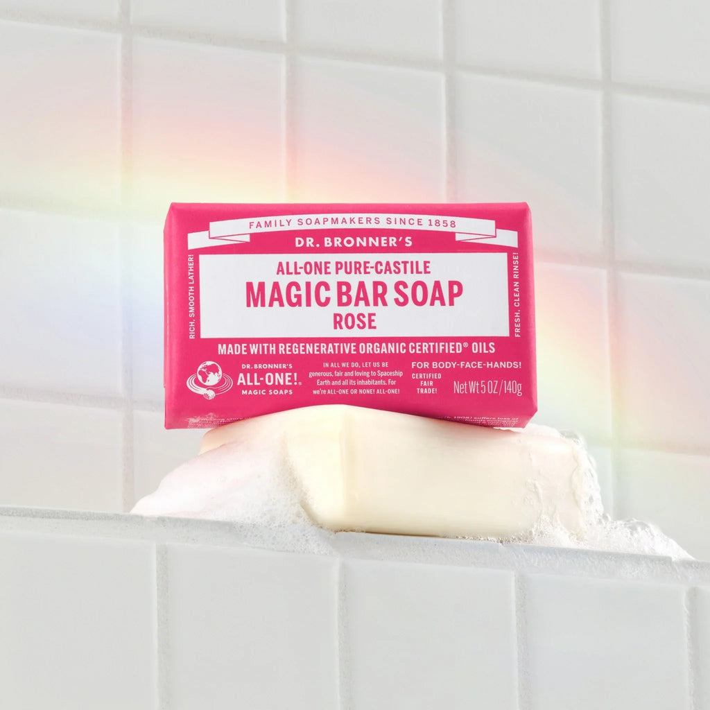 A Dr. Bronners Rose Magic Bar Soap rests on a tiled surface with foam. The packaging reads DR. BRONNERS ALL ONE PURE CASTILE MAGIC BAR SOAP ROSE, emphasizing regenerative organic certified oils. A subtle rainbow appears over the soap [5 oz]
