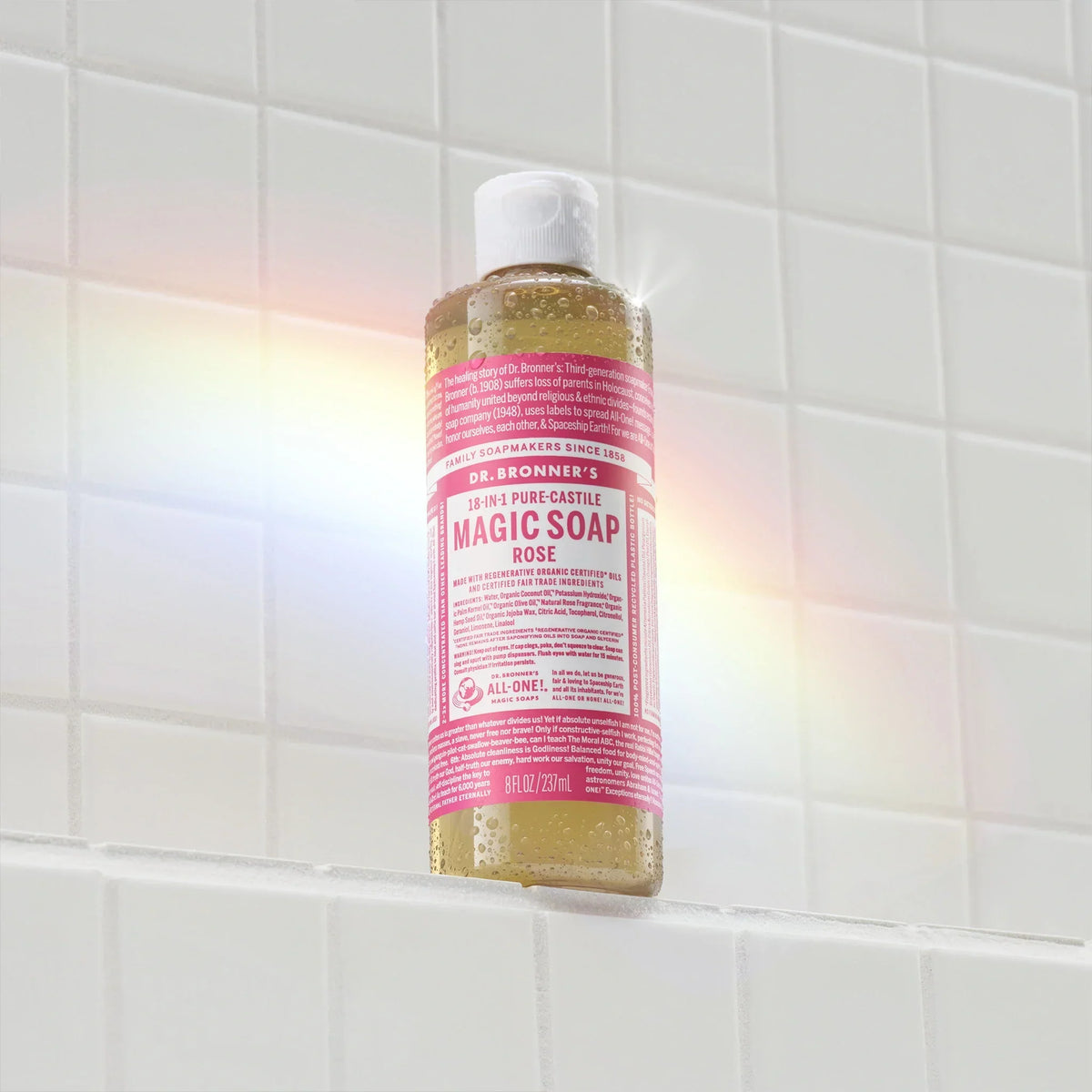 A bottle of Dr. Bronners 18 in 1 Pure Castile Magic Soap Rose rests against white tiles, illuminated by a colorful light reflection. Text reads Dr. Bronners 18 in 1 Pure Castile Magic Soap Rose, 8 fl oz 237 mL [8 oz]
