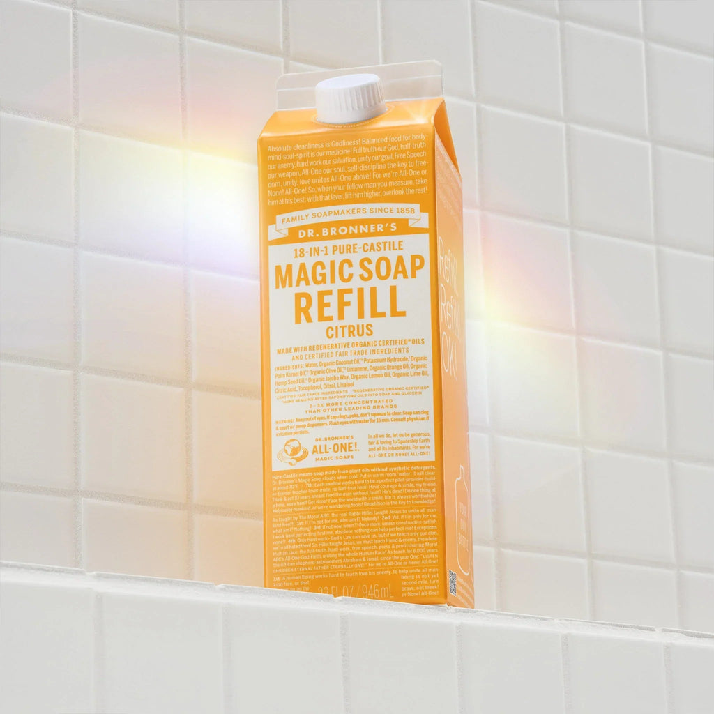 An orange liquid soap refill carton labeled Dr. Bronners MAGIC SOAP REFILL stands upright on a white tiled surface. Sunlight creates subtle rainbow reflections around it, enhancing the eco friendly packagings appeal [32 oz]