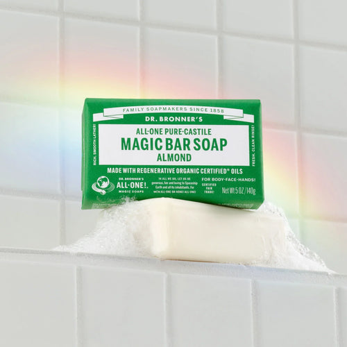 Dr. Bronners Magic Bar Soap in almond scent sits on a soap dish, generating bubbles against a tiled wall with a visible rainbow reflection. FAMILY SOAPMAKERS SINCE 1858, ALL ONE PURE CASTILE MAGIC BAR SOAP ALMOND, MADE WITH REGENERATIVE ORGANIC CERTIFIED OILS, FOR BODY FACE HANDS CERTIFIED FAIR TRADE [5 oz]