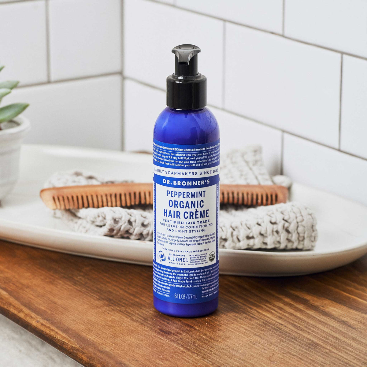 A blue bottle of Dr. Bronners Peppermint Organic Hair Crme sits on a wooden countertop. Nearby, a comb and folded towels create a bathroom setting, enhanced by a tiled wall backdrop [6 oz]