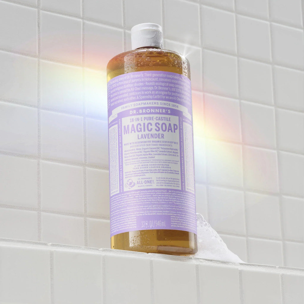 Dr. Bronner’s Lavender 18-in-1 Pure Castile Magic Soap bottle on white tiles surrounded by soap suds with a faint rainbow reflection.