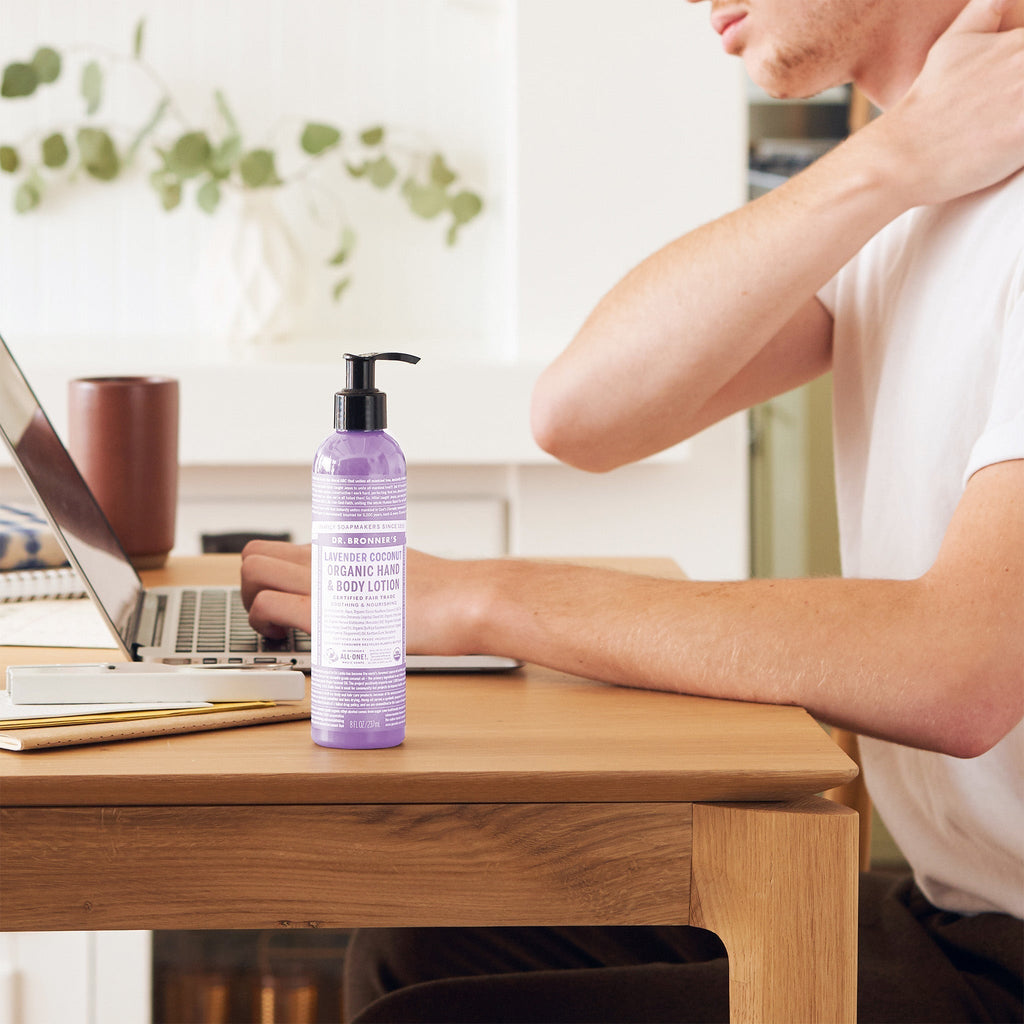 A bottle of Dr. Bronners Lavender Organic Hand Body Lotion sits on a wooden desk beside a person using a laptop, with notebooks and a plant in the background [8 oz]