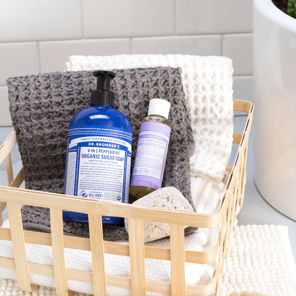 A bamboo basket holds Dr. Bronners 4 in 1 Peppermint Organic Sugar Soap and 18 in 1 Hemp Lavender Pure Castile Soap, atop textured towels beside a white tiled wall. DR. BRONNERS 4 IN 1 PEPPERMINT ORGANIC SUGAR SOAP DR. BRONNERS 18 IN 1 HEMP LAVENDER PURE CASTILE SOAP [4 oz]