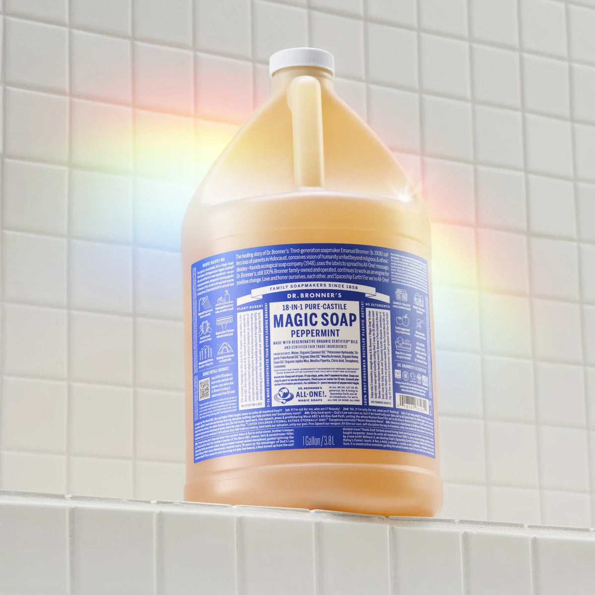 A large bottle of Dr. Bronners 18 in 1 Pure Castile Magic Soap, Peppermint, sits on white tiled surface, surrounded by a subtle rainbow reflection. Text reads 1 Gallon 3.8L [1 Gallon]
