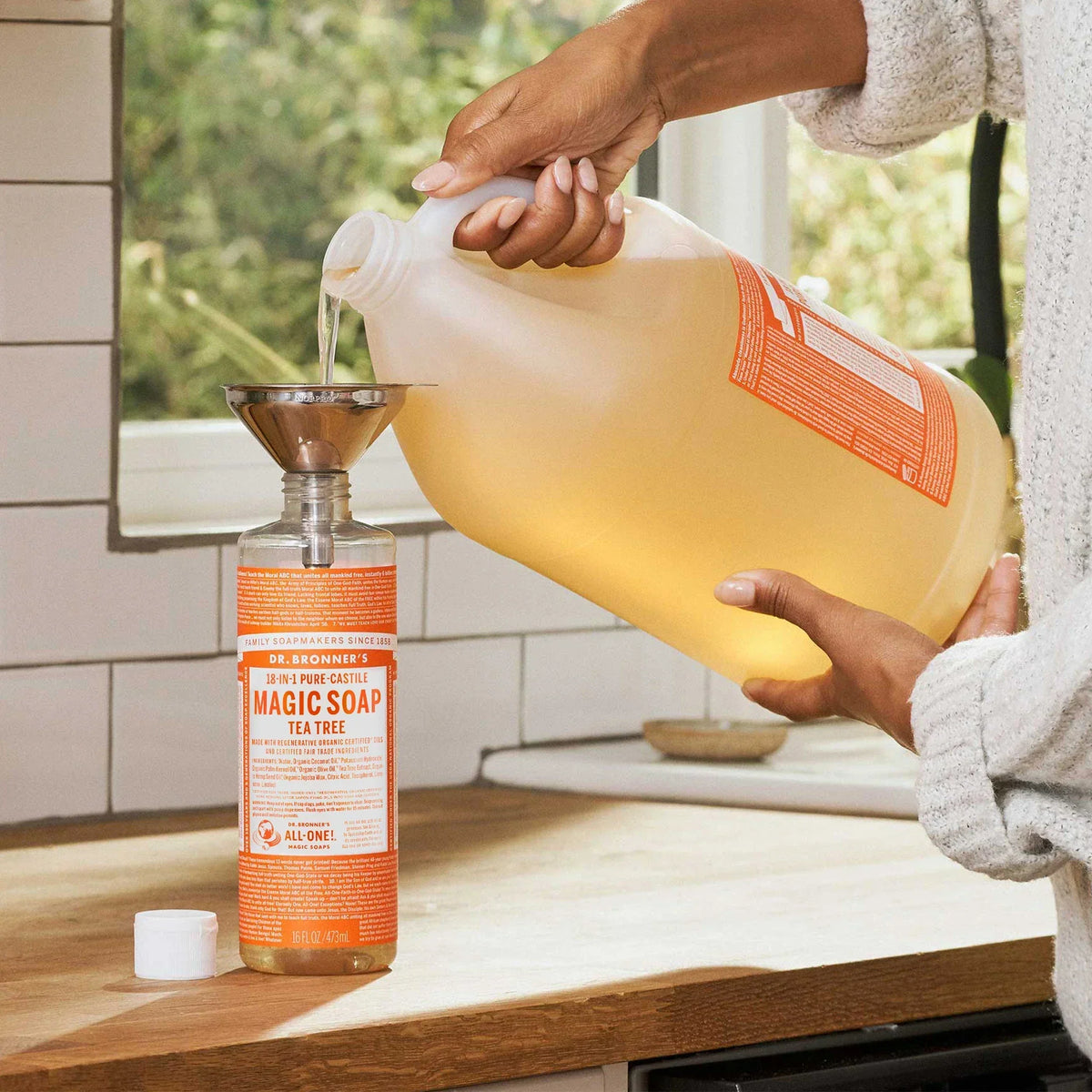 A large bottle of Dr. Bronners Tea Tree Pure Castile Liquid Soap is being poured into a smaller bottle using a funnel on a kitchen counter. Sunlit greenery is visible through a window [16 oz]