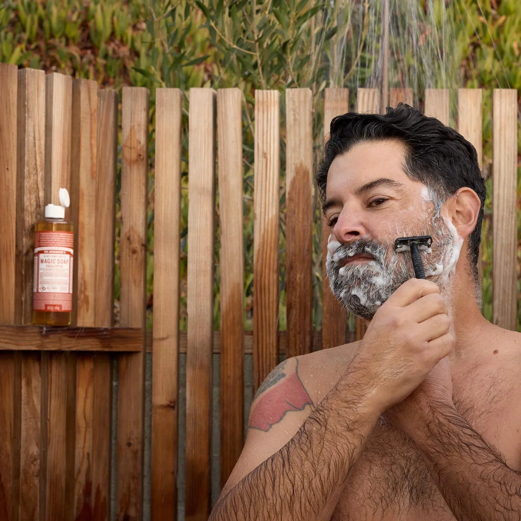 A man shaves outdoors using a razor and Dr Bronners soap, surrounded by a wooden fence and greenery. A bottle of Dr Bronners organic soap rests on a shelf nearby, enhancing the natural setting [8 oz]