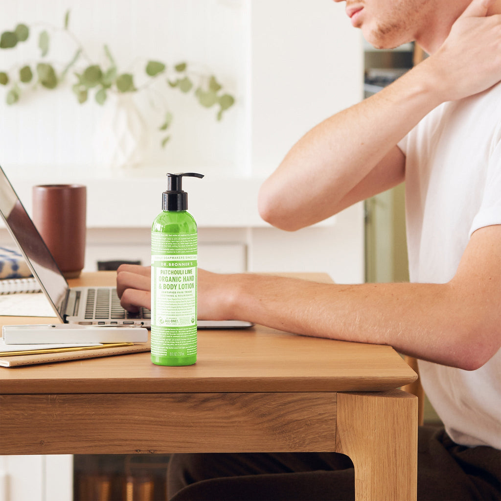 A green bottle of Dr. Bronners Patchouli Lime Organic Hand Body Lotion sits on a wooden table. A person uses a laptop in a cozy, plant adorned room, touching their neck [8 oz]