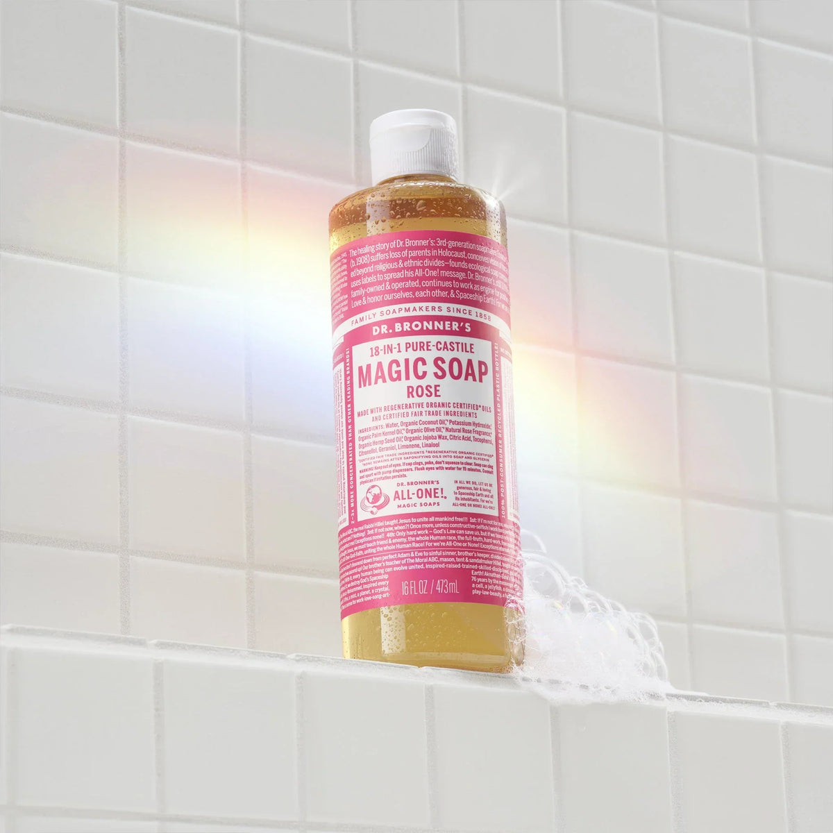 A bottle of Dr. Bronners Rose Castile Soap stands upright on a white tiled surface. A subtle rainbow reflection graces the scene, adding a vibrant touch to the minimalist bathroom setting  [16 oz]