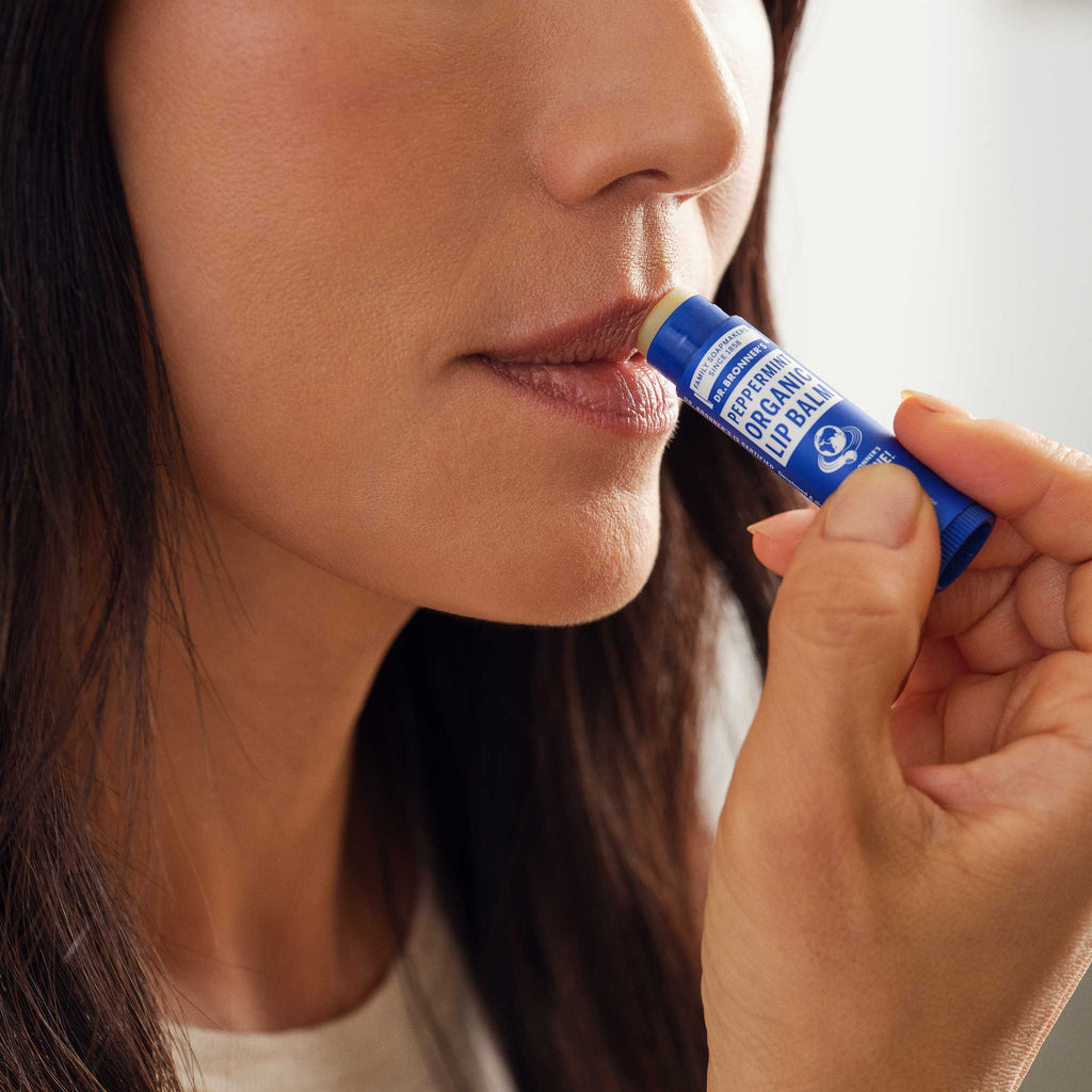 A person applies Dr Bronners Peppermint Organic Lip Balm to their lips, highlighting natural care with organic, eco friendly ingredients. The context suggests a focus on personal wellness and sustainability [.15 oz]