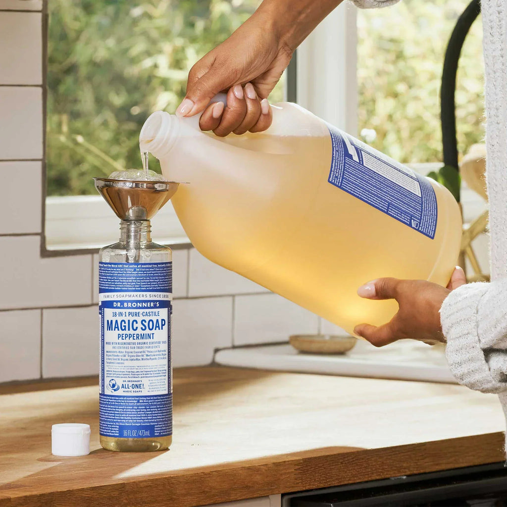 A large jug of liquid is being poured into a bottle through a funnel on a wooden kitchen counter. The visible small bottle is labeled Dr. Bronners Pure Castile Liquid Soap, surrounded by white subway tiles and a window showing greenery [16 oz]
