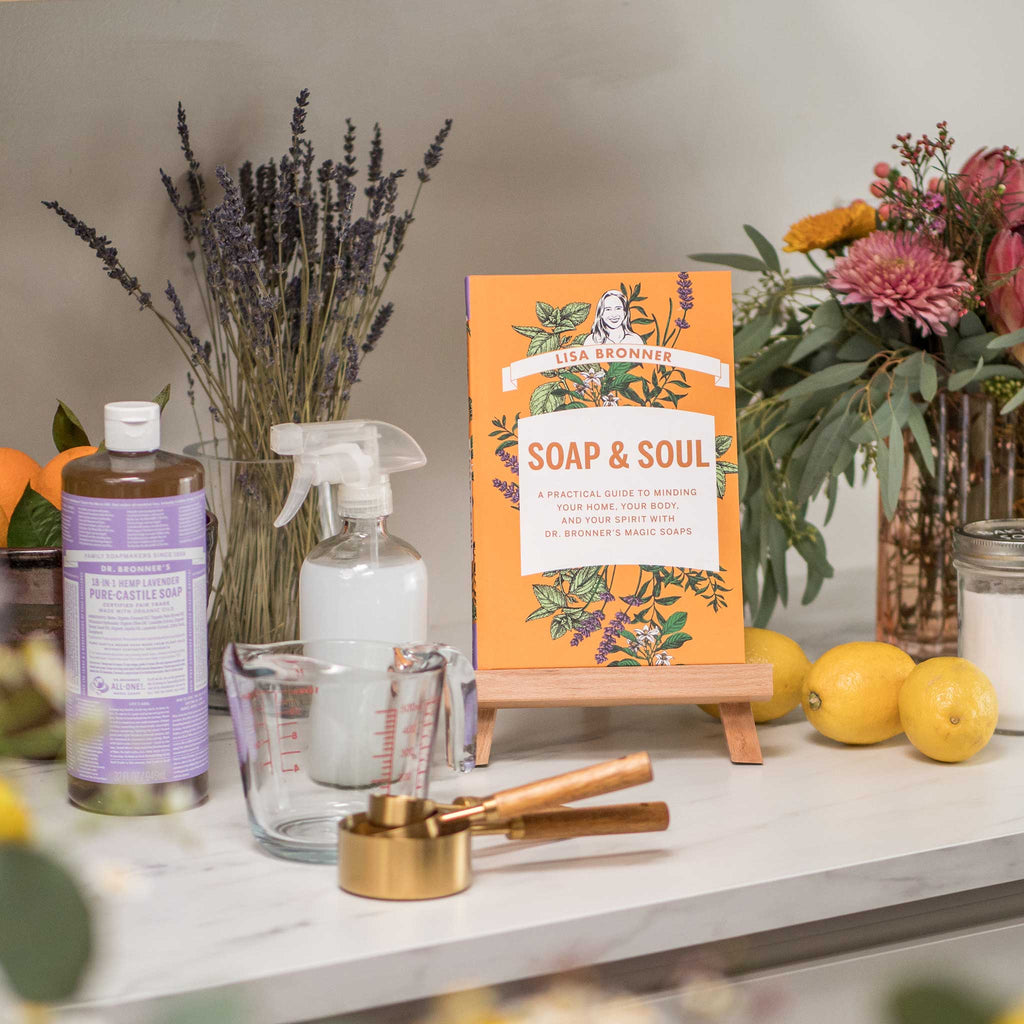 A bottle of Dr. Bronners lavender pure castile soap stands beside a spray bottle, measuring cup, and golden measuring spoons on a countertop. An orange book titled Lisa Bronner Soap Soul is displayed on a wooden stand
