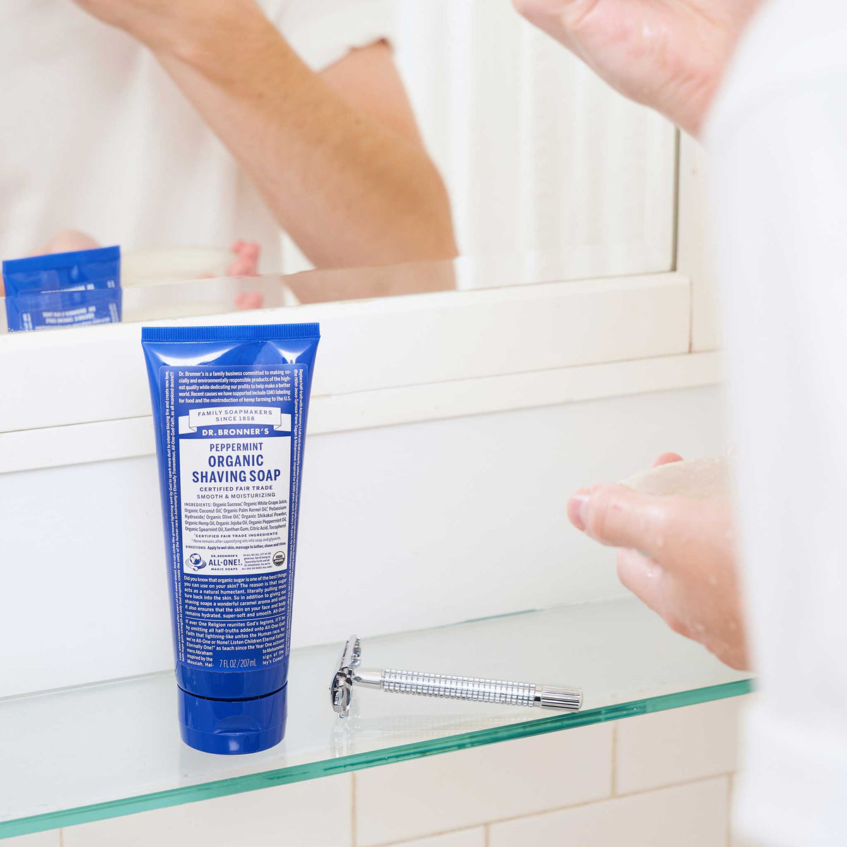 A tube of Dr. Bronners Peppermint Organic Shaving Soap sits on a bathroom glass shelf, accompanied by a razor. A person is using the soap, reflected in the mirror [7 oz]
