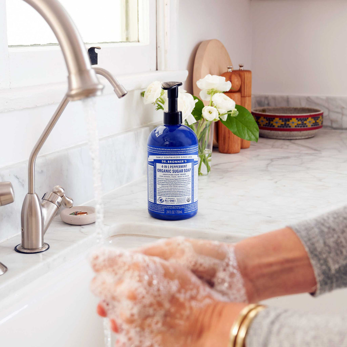 A person washes hands with foamy soap in a kitchen sink. Dr. Bronners 4 in 1 Peppermint Organic Sugar Soap sits on the counter beside flowers and kitchen items [24 oz]